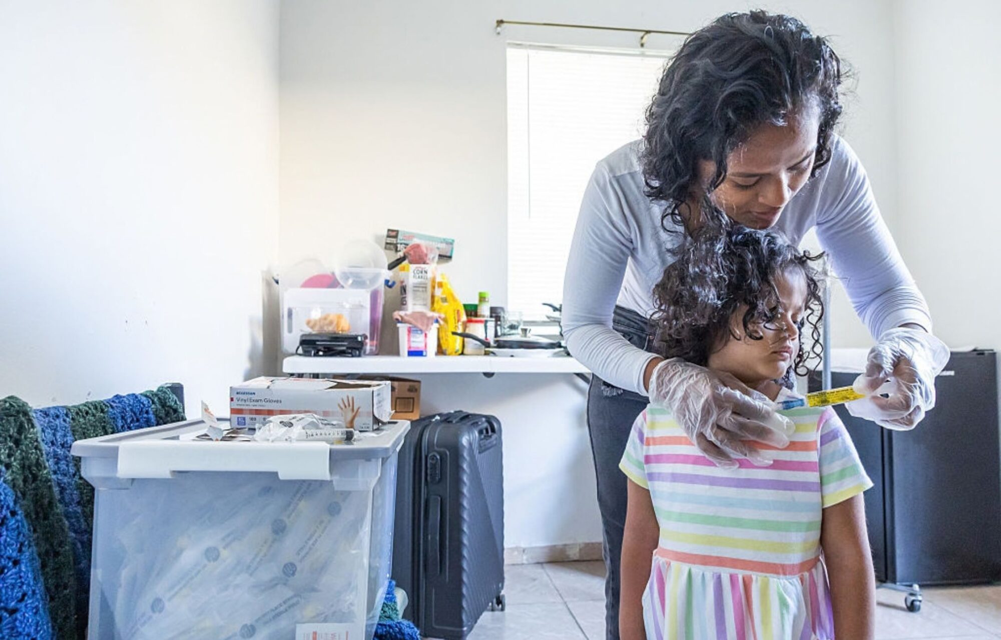 A woman with curly black hair is giving her four-year-old child, wearing a rainbow-colored dress, her daily flush solution through her intravenous line.