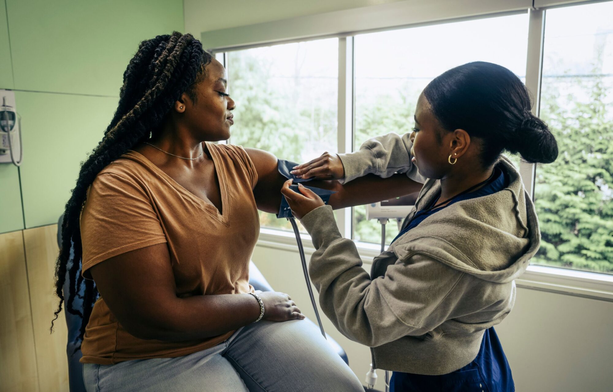 A black female medical professional with straight hair pulled into a bun is wrapping a blood pressure cuff around the arm of a black female patient with long braids