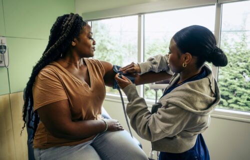 A black female medical professional with straight hair pulled into a bun is wrapping a blood pressure cuff around the arm of a black female patient with long braids
